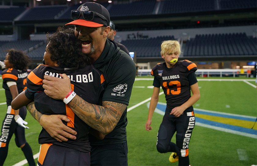 Youth football coach hugging a player during a team celebration on a stadium field.